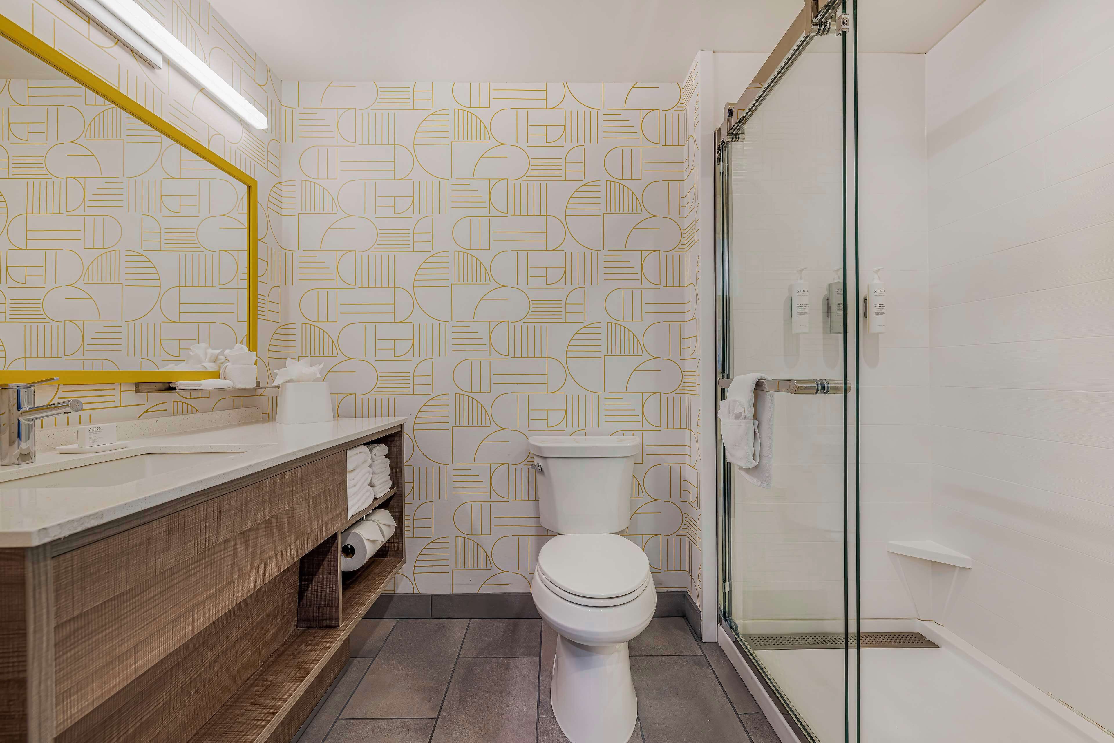A modern bathroom with a geometric patterned wall, a white toilet, a glass shower enclosure, and a wooden vanity with towels.