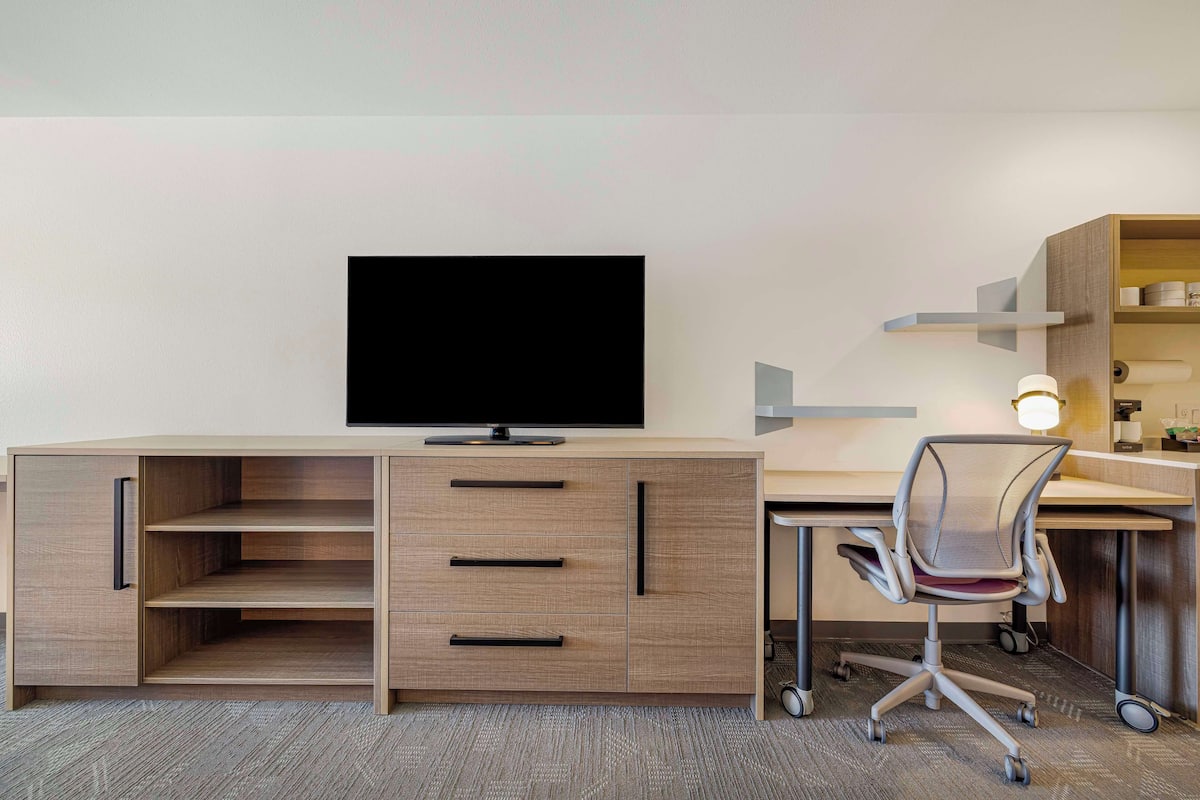 Un espace de bureau moderne, doté d’un téléviseur à écran plat fixé sur un meuble en bois, d’un bureau avec une chaise et d’une étagère contenant divers objets.