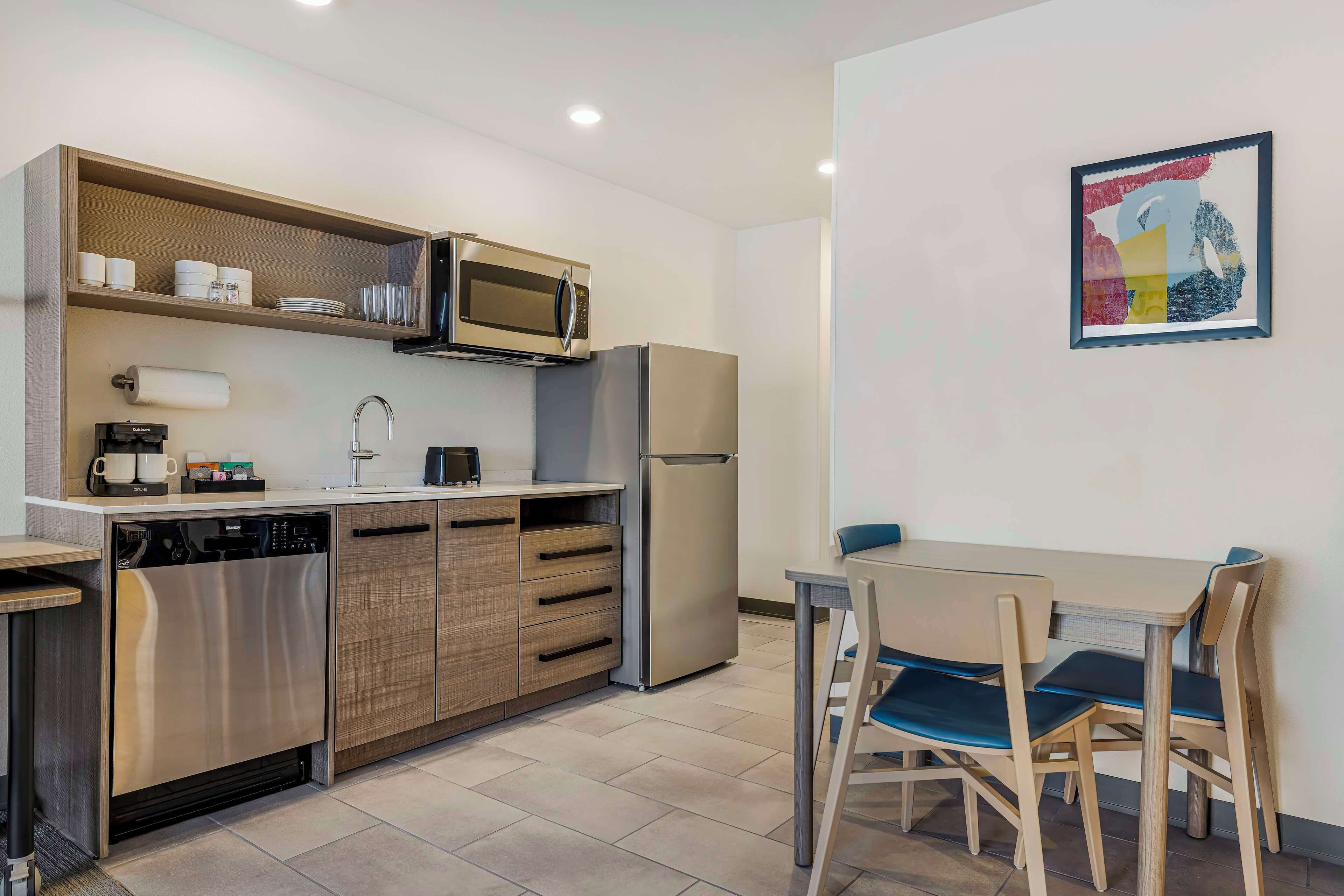 A compact kitchen with a microwave, refrigerator, and dishwasher, a small dining area with two chairs, and a wall-mounted artwork.