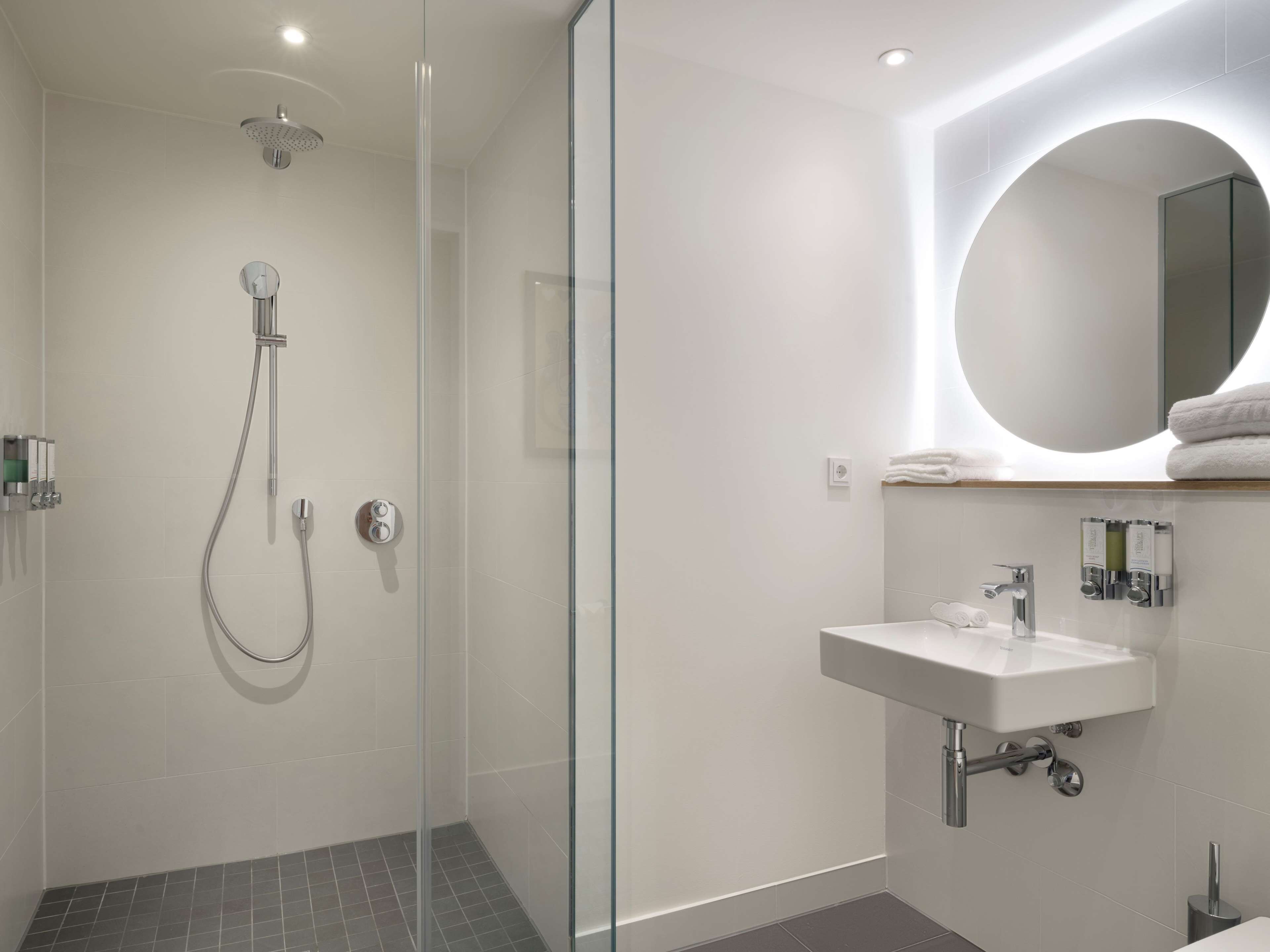 A modern bathroom with a glass-enclosed shower, a white sink, and a round mirror.