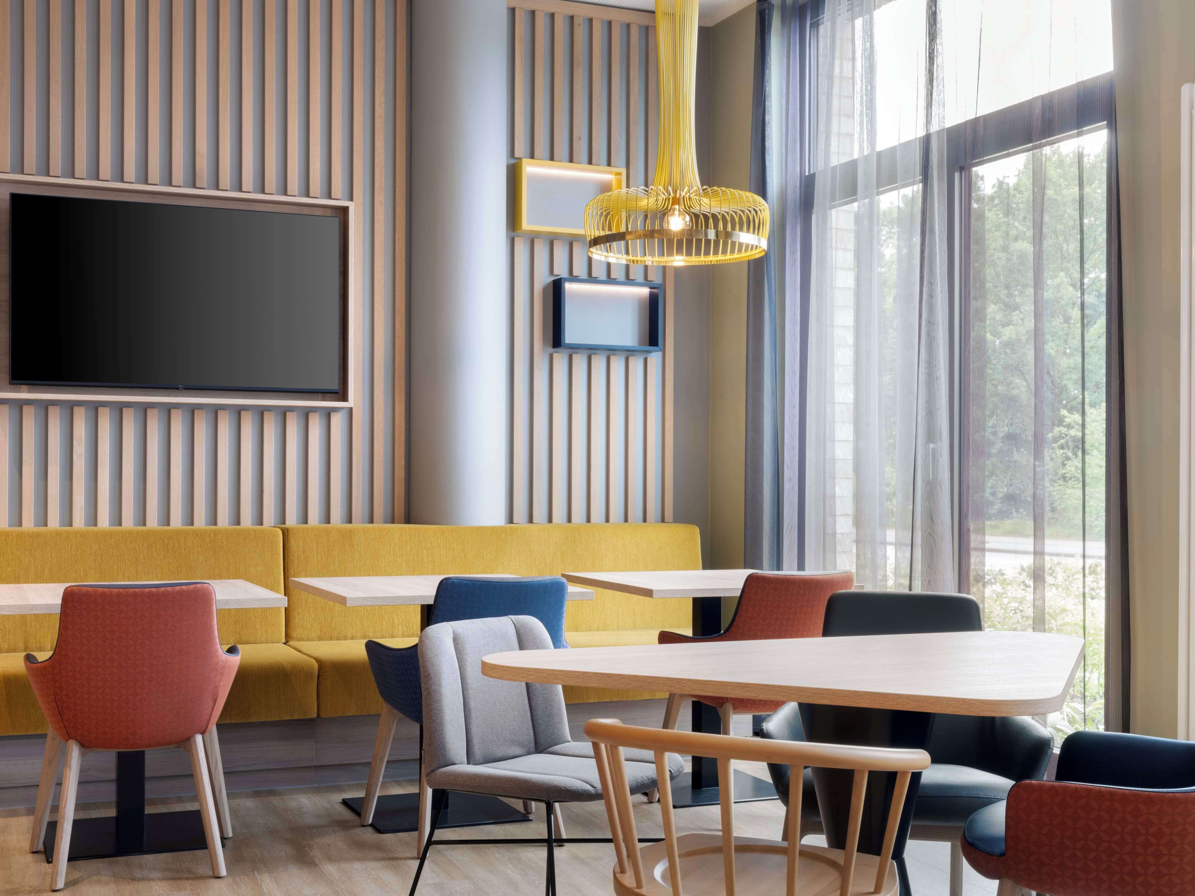 A modern dining area with a yellow booth seating, a large TV mounted on the wall, and a large window with a view of trees.