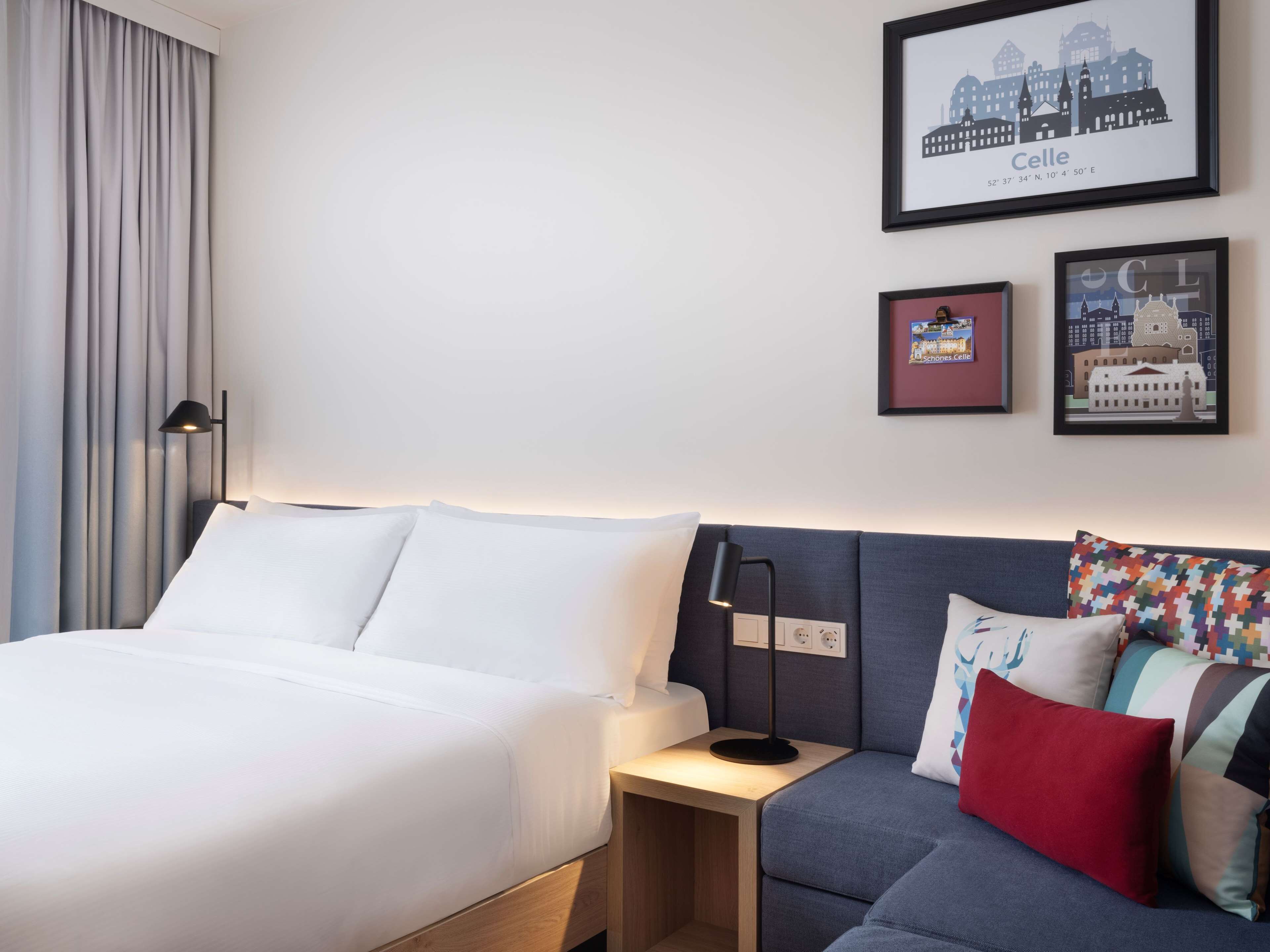 Ein Hotelzimmer mit Bett, Sofa und Nachttisch mit Lampe. An der Wand hängen gerahmte Bilder.