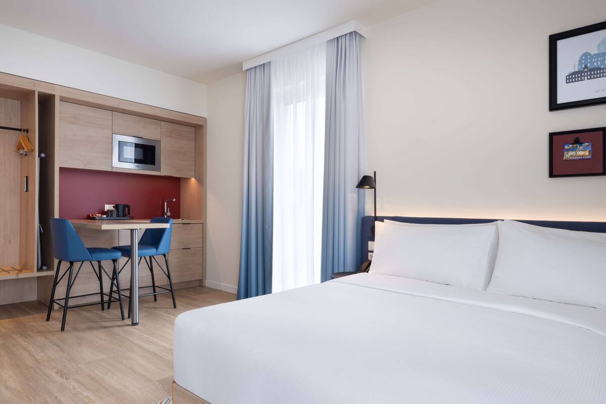A hotel room with a bed, a desk with two chairs, a microwave, and a small kitchenette.