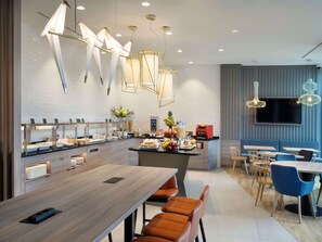 A modern restaurant with a long communal table, individual seating, a buffet area, and a flat-screen TV mounted on the wall. - Hampton By Hilton Celle (Celle)
