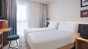A hotel room with two beds, a desk, a chair, a TV, and a large window with curtains. - Hampton By Hilton Celle (Celle)