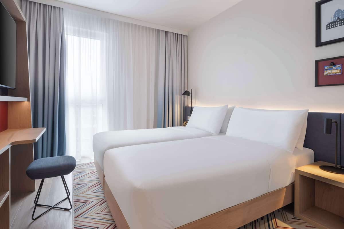 A hotel room with two beds, a desk, a chair, a TV, and a large window with curtains.