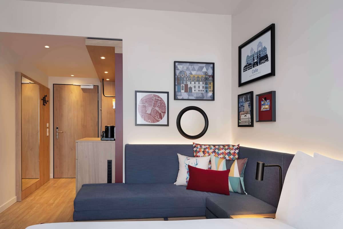 A modern hotel room with a grey sofa, a bed, and a small kitchenette. There are framed pictures on the wall and a mirror above the sofa.