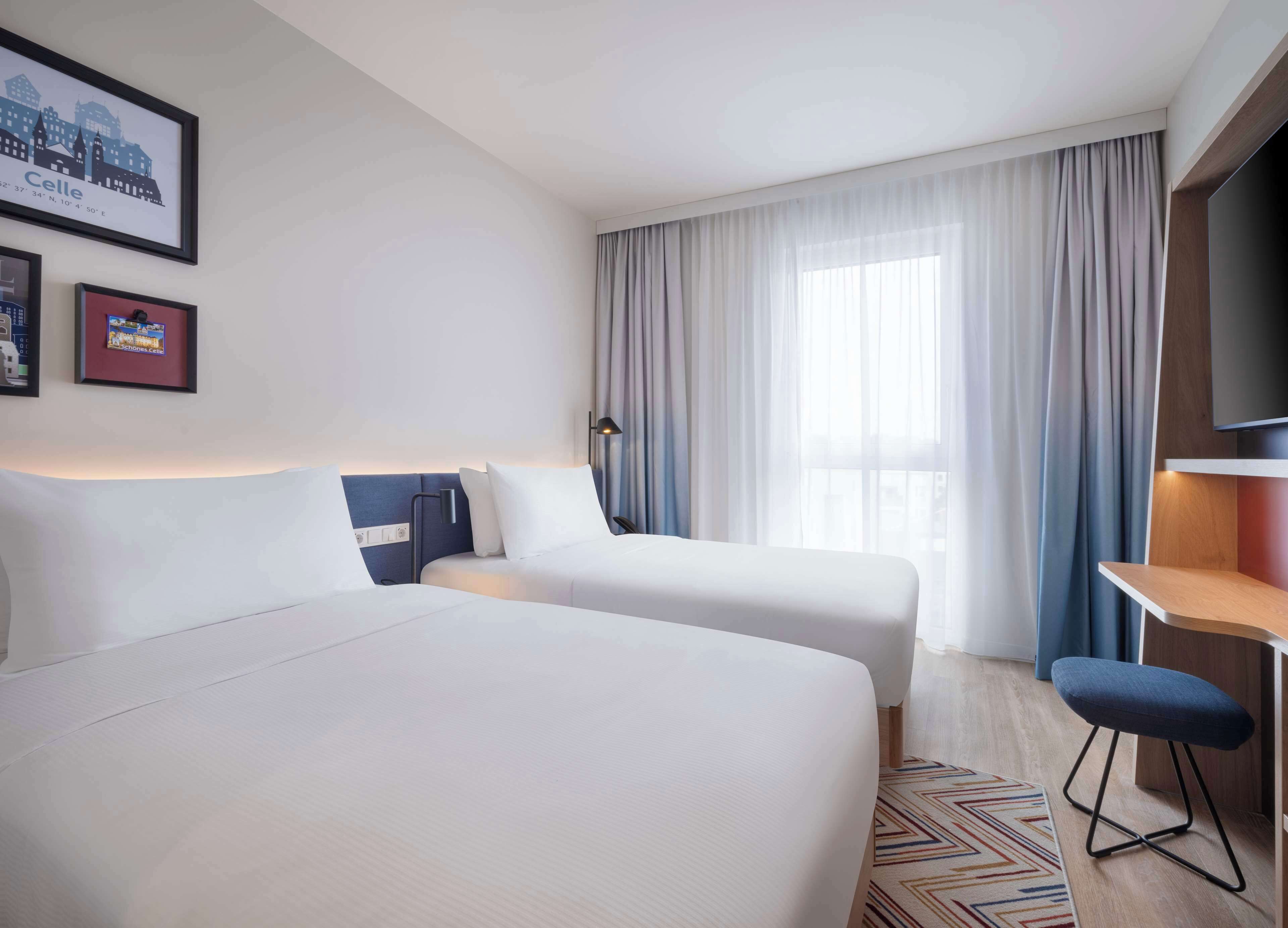 A hotel room with two beds, a desk, a TV, and a window with curtains.