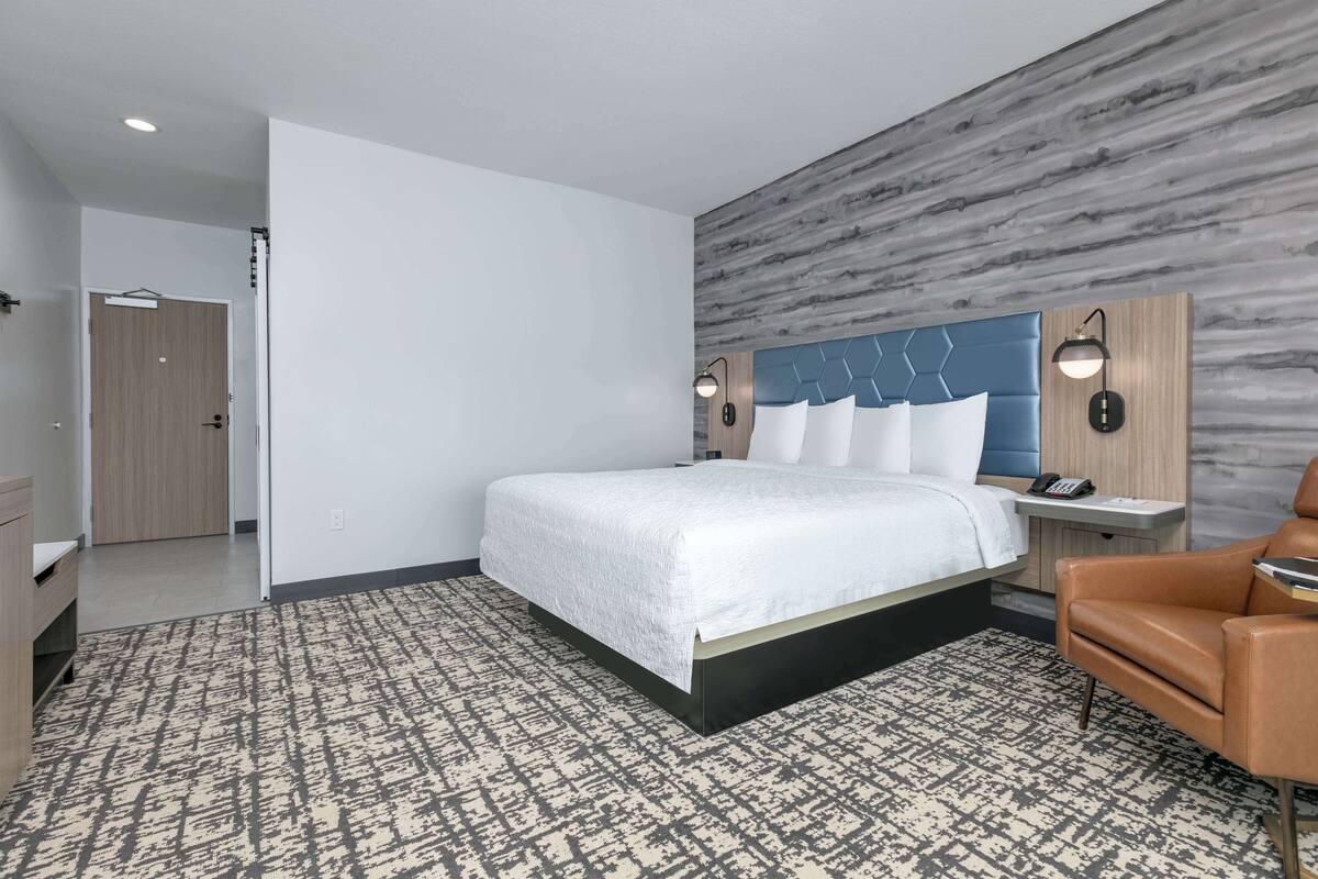 A modern hotel room with a large bed, a desk, a chair, and a patterned carpet.