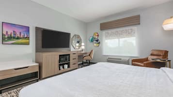 Quarto de hotel com uma cama grande, uma televisão de tela plana, uma escrivaninha de madeira com uma luminária, uma poltrona de couro marrom e uma pintura de paisagem urbana na parede.
