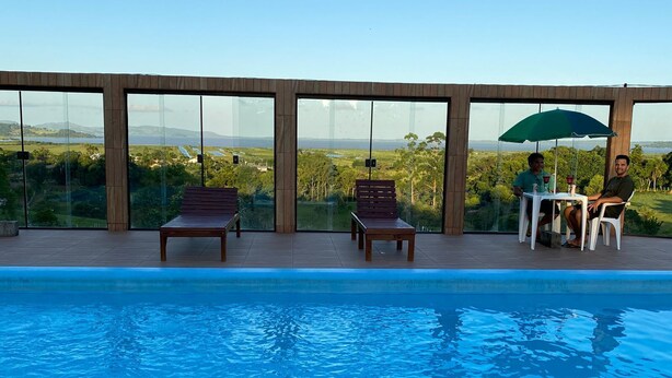 Área da piscina com duas espreguiçadeiras de madeira e uma mesa com duas pessoas sob um guarda-sol verde.