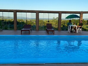 A pool area with two wooden lounge chairs and a table with two people under a green umbrella. - POUSADA SITIO TRES LAGOS (Pescaria Brava)