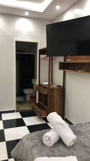 A bathroom with a black and white checkered floor, a wooden vanity with a mirror, a television mounted above it, and a towel on a gray mat. - POUSADA SITIO TRES LAGOS (Pescaria Brava)