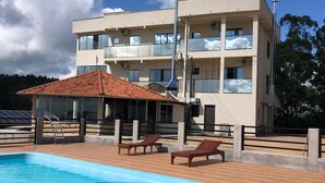Piscina com espreguiçadeiras, área coberta e prédio com várias andares e varandas.