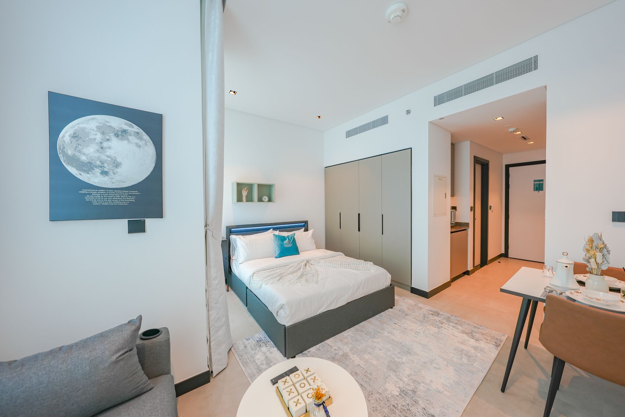 A modern hotel room with a bed, a sofa, a dining table, and a kitchen area.