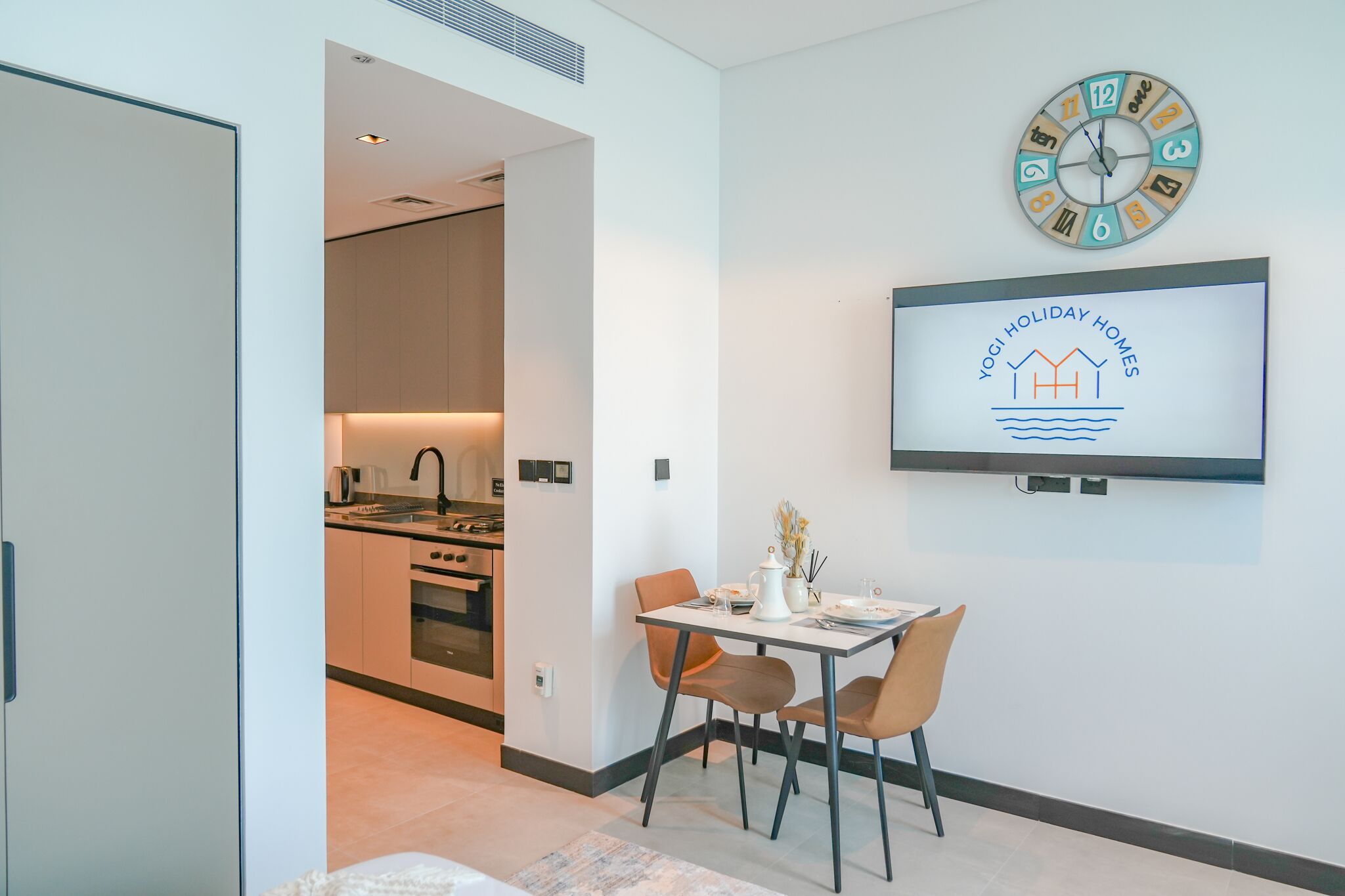 A modern kitchen with a dining area, a clock, and a TV displaying 'Yogi Holiday Homes'.