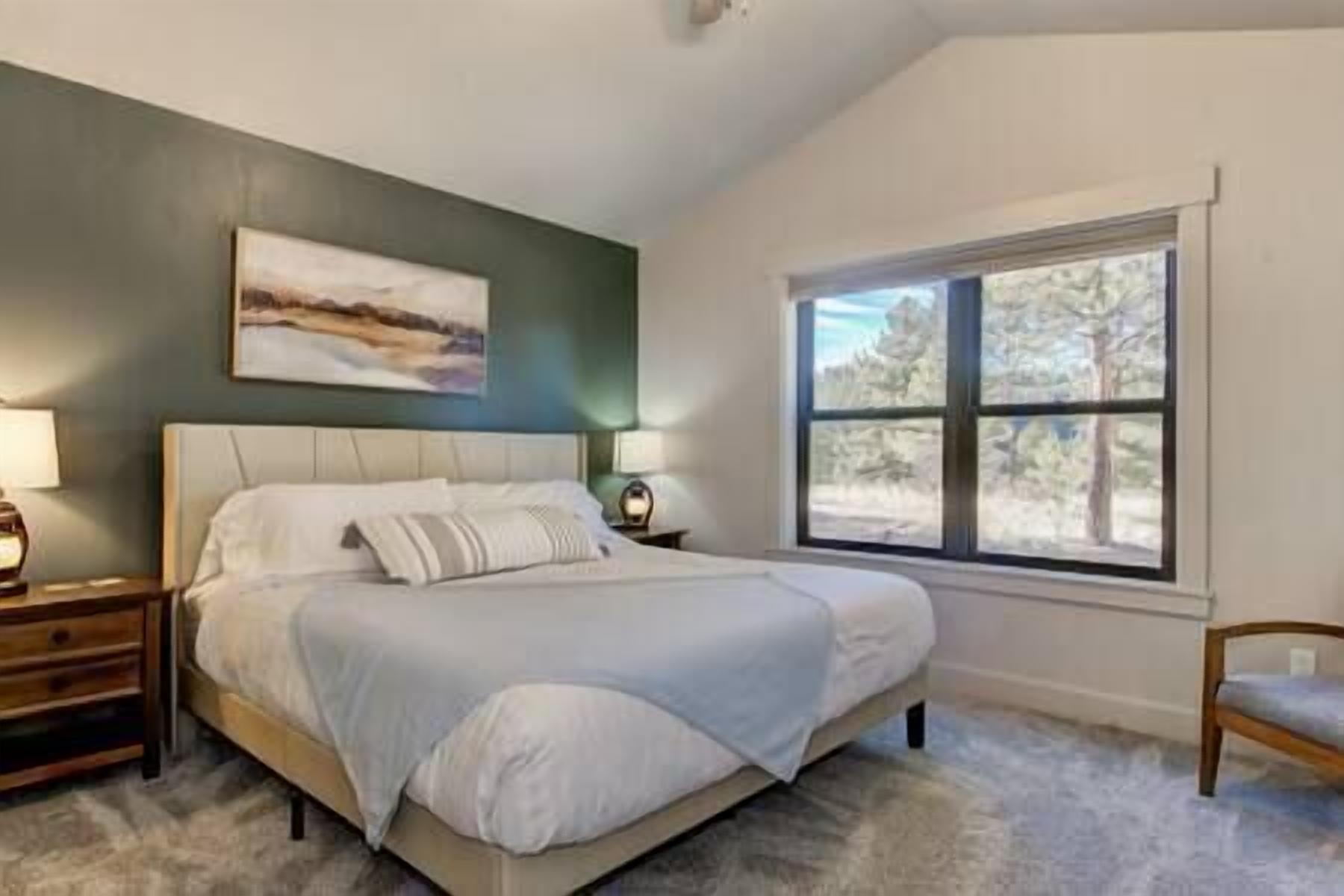 A bedroom with a large bed, a nightstand, a lamp, a chair, and a window with a view of trees.