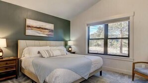 Une chambre à coucher avec un grand lit, une table de chevet, une lampe, une chaise et une fenêtre donnant sur des arbres.