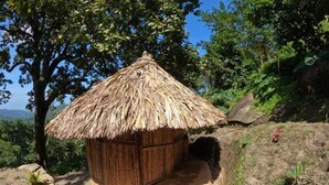 A small wooden hut with a thatched roof, surrounded by lush greenery and a rocky landscape. - Private waterfall and indigenous house! (Minca)