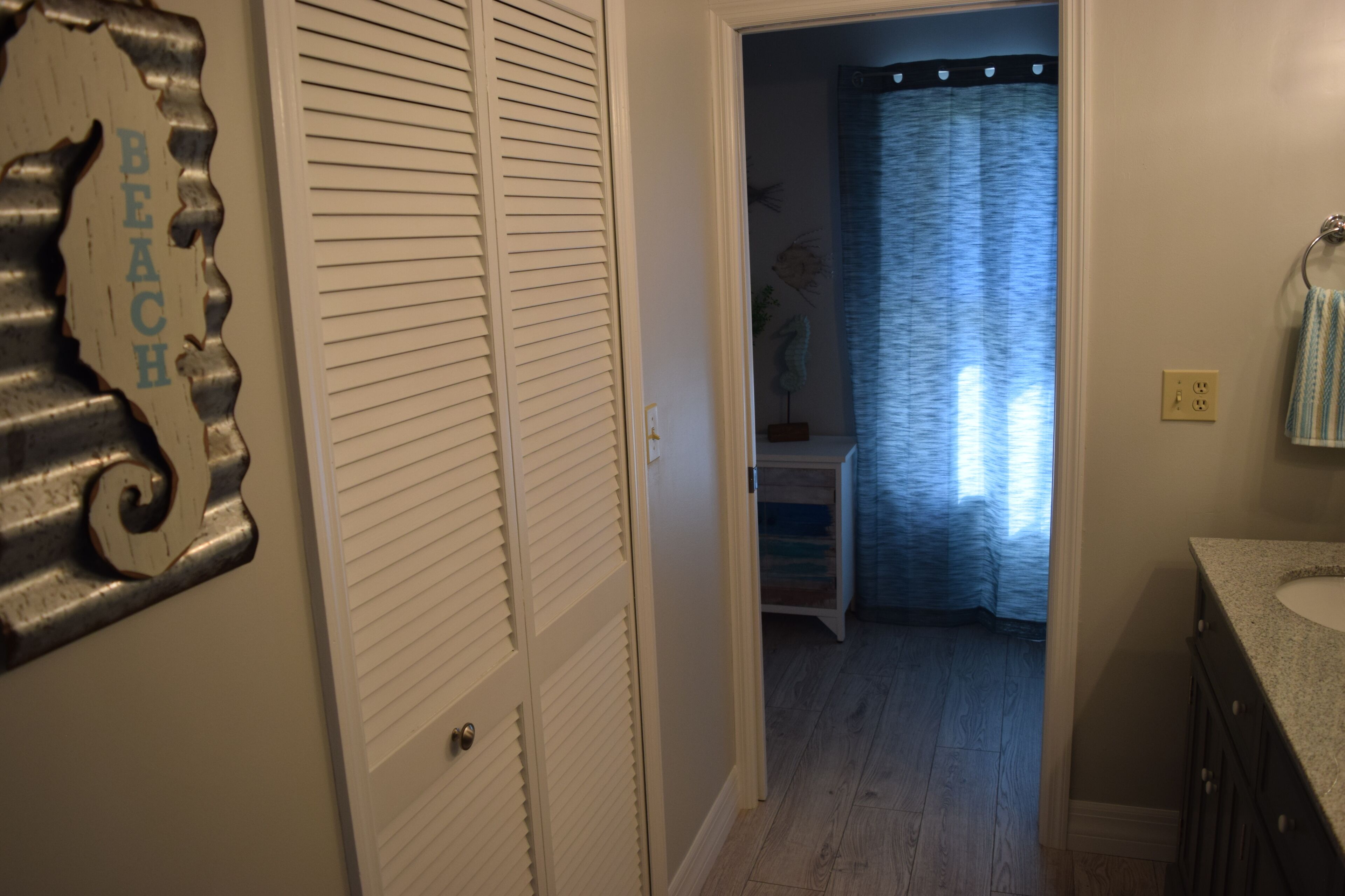 A hallway with a white door, a metal sign reading "BEACH", a small table with a blue curtain, and a bathroom with a sink and towel rack.