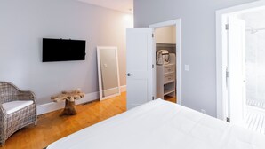 A bedroom with a bed, a television mounted on the wall, a wicker chair, a wooden table, and a wardrobe.