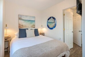 Un dormitorio con una cama, dos mesitas de noche, un cuadro colgado en la pared, un espejo circular y una puerta.