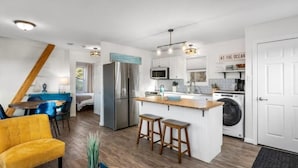 Una cocina compacta con una isla central, un comedor con una silla azul y un sofá amarillo.