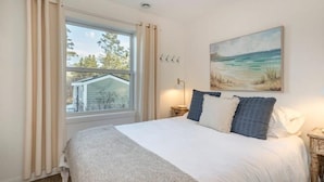 Un dormitorio con una cama grande, un cuadro de una playa, una ventana con cortinas y una mesita de noche con una lámpara.