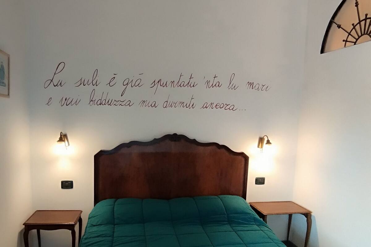 Quarto com colcha verde-água, cabeceira de madeira, duas mesas de cabeceira com luminárias e parede com frase decorativa.