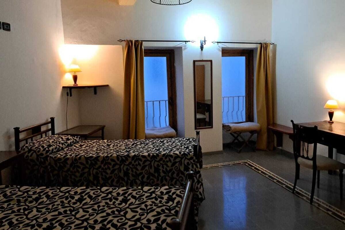 Quarto de hotel com duas camas, uma escrivaninha, uma cadeira, uma mesinha e varanda com cortinas.