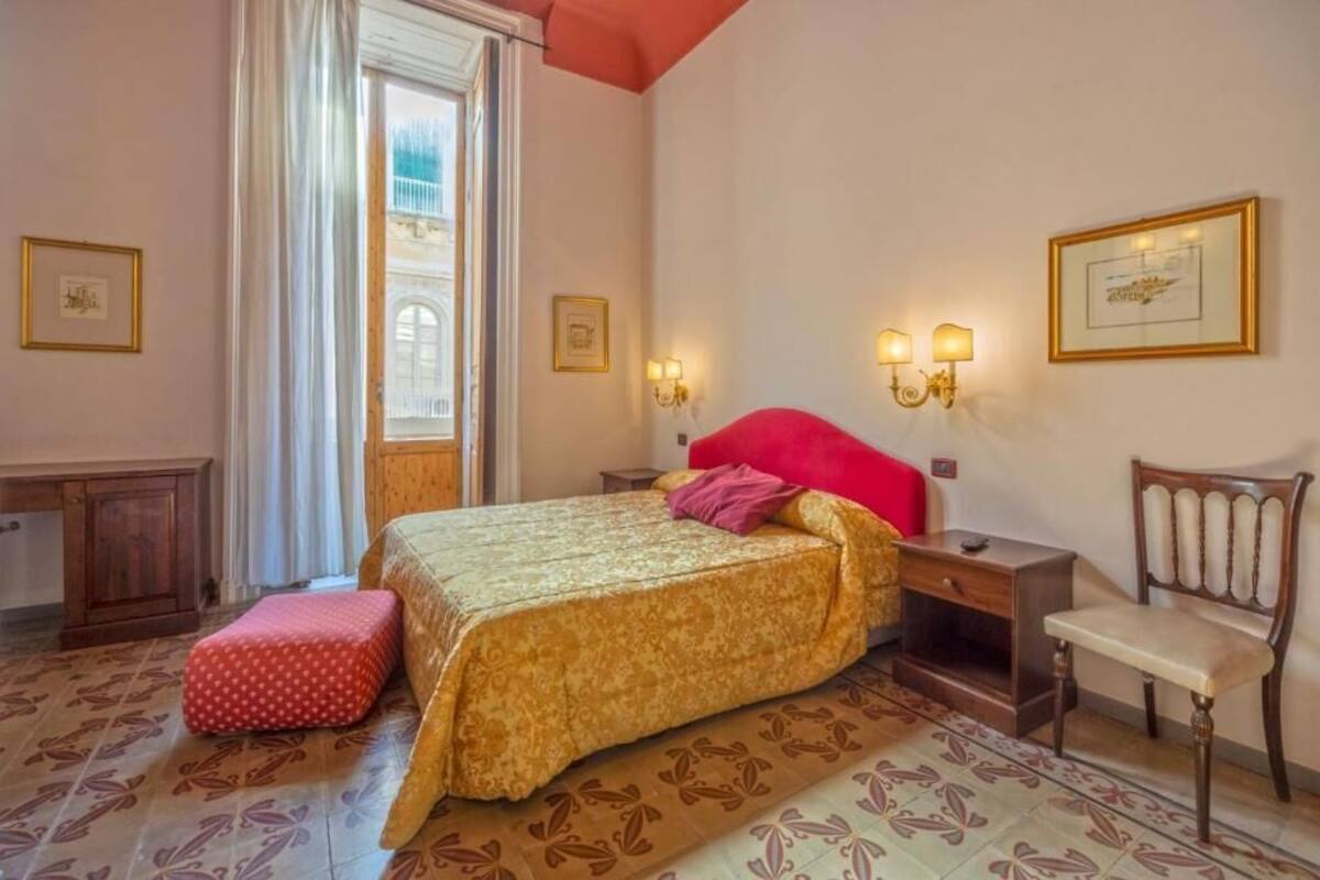 Um quarto com cama, cadeira, escrivaninha e janela.
