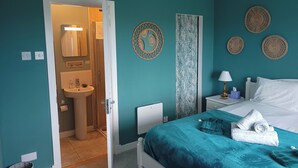 A bedroom with teal walls, a bed with a teal bedspread, a white headboard, a nightstand with a lamp, and a wall-mounted shelf. - Fishermans Holding (Isle of Lewis)