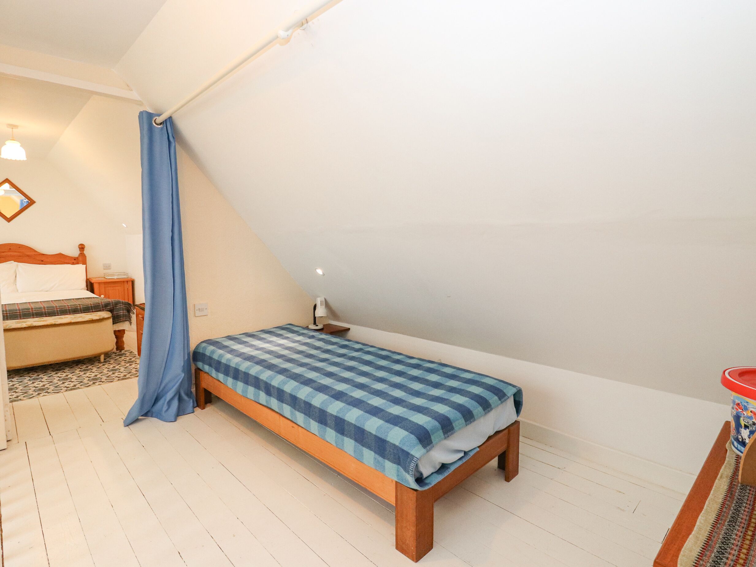 A small, single-bed room with a sloped ceiling, a wooden bed frame, a blue checkered bedspread, a nightstand with a lamp, and a small table with a red container.
