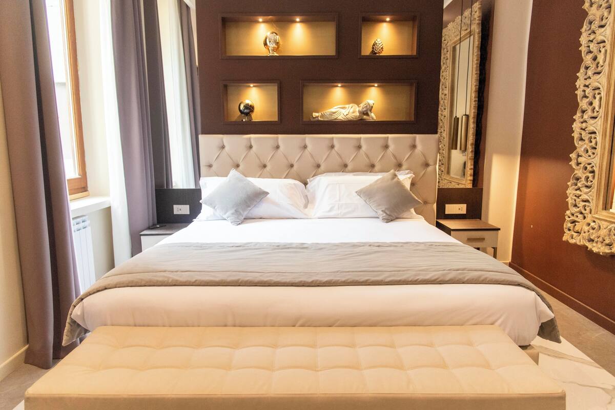 A bedroom with a large bed, a headboard, bedside tables, a nightstand, a mirror, and wall-mounted shelves with decorative items.