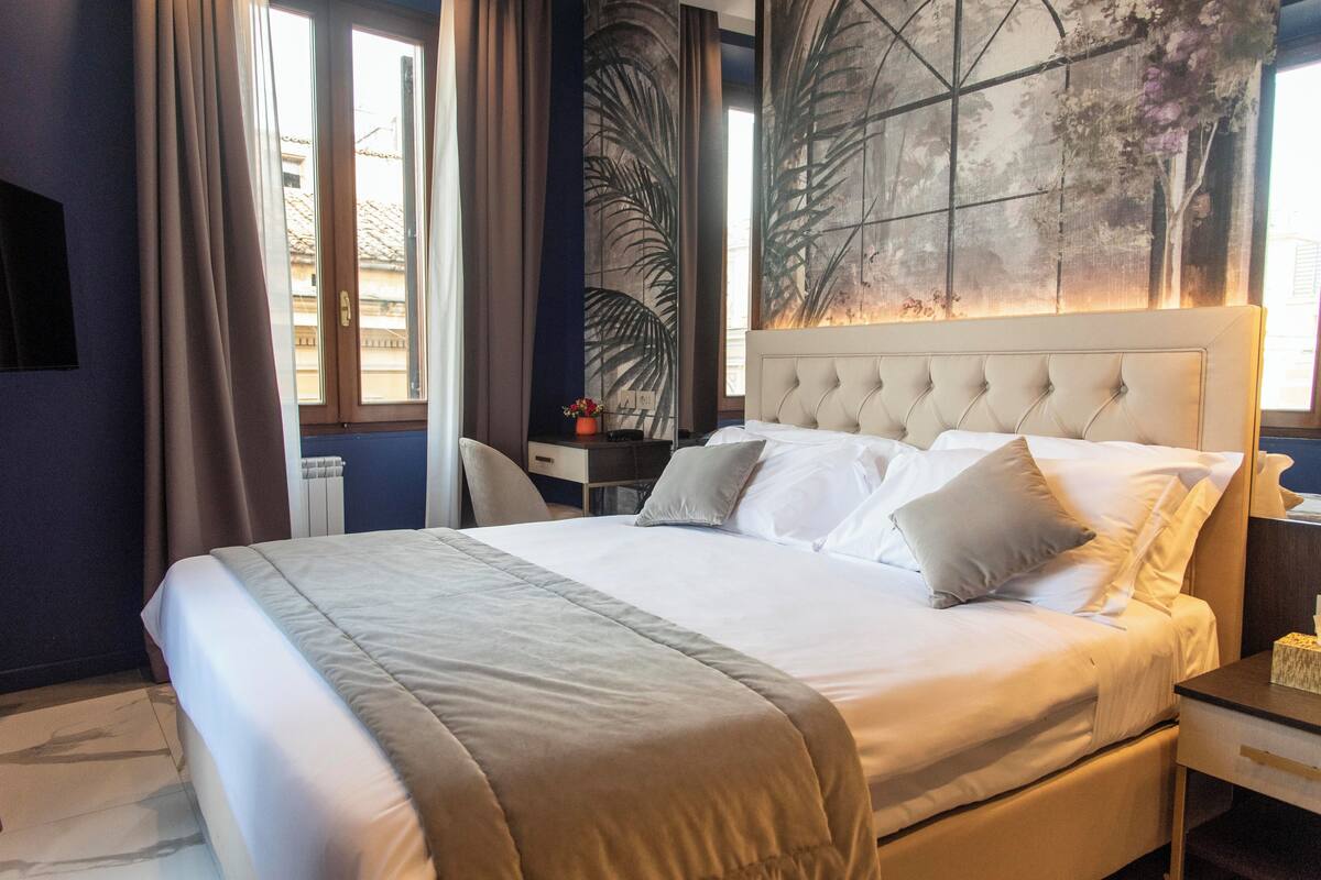 A neatly made bed with a tufted headboard, a bedside table with a lamp, and a large window with curtains.