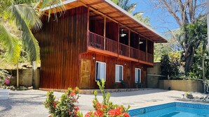 A wooden two-story building with a balcony, a swimming pool with blue tiles, and potted plants with red flowers. - TREE HOUSE PLAYA SAMARA (Playa Samara Guanacaste Province)