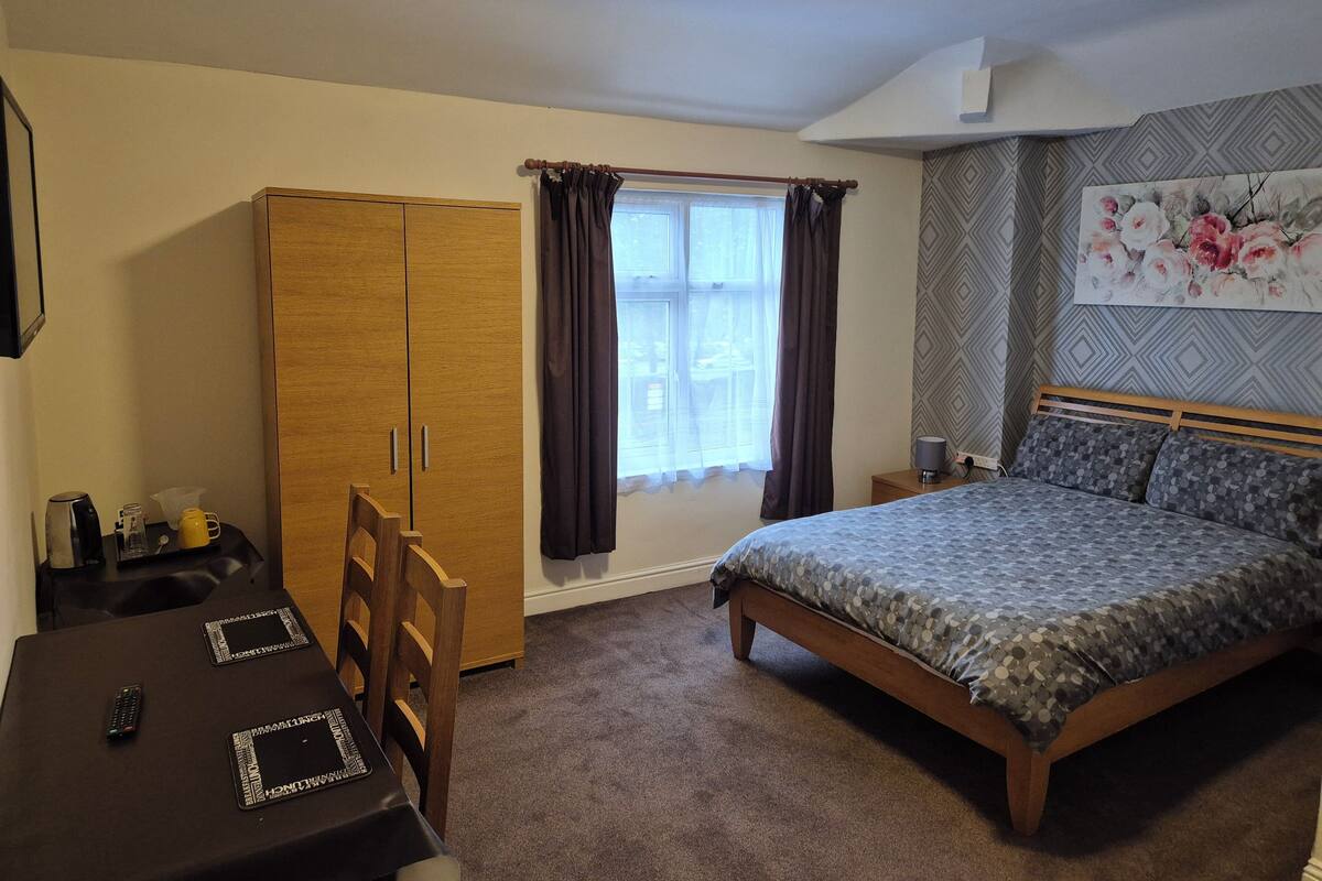 A bedroom with a bed, a wooden wardrobe, a table with a remote control, and a window with curtains.
