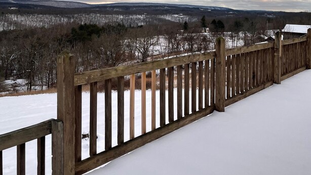 Une terrasse en bois recouverte de neige, d’où l’on peut admirer un paysage enneigé avec des arbres et des bâtiments éloignés.