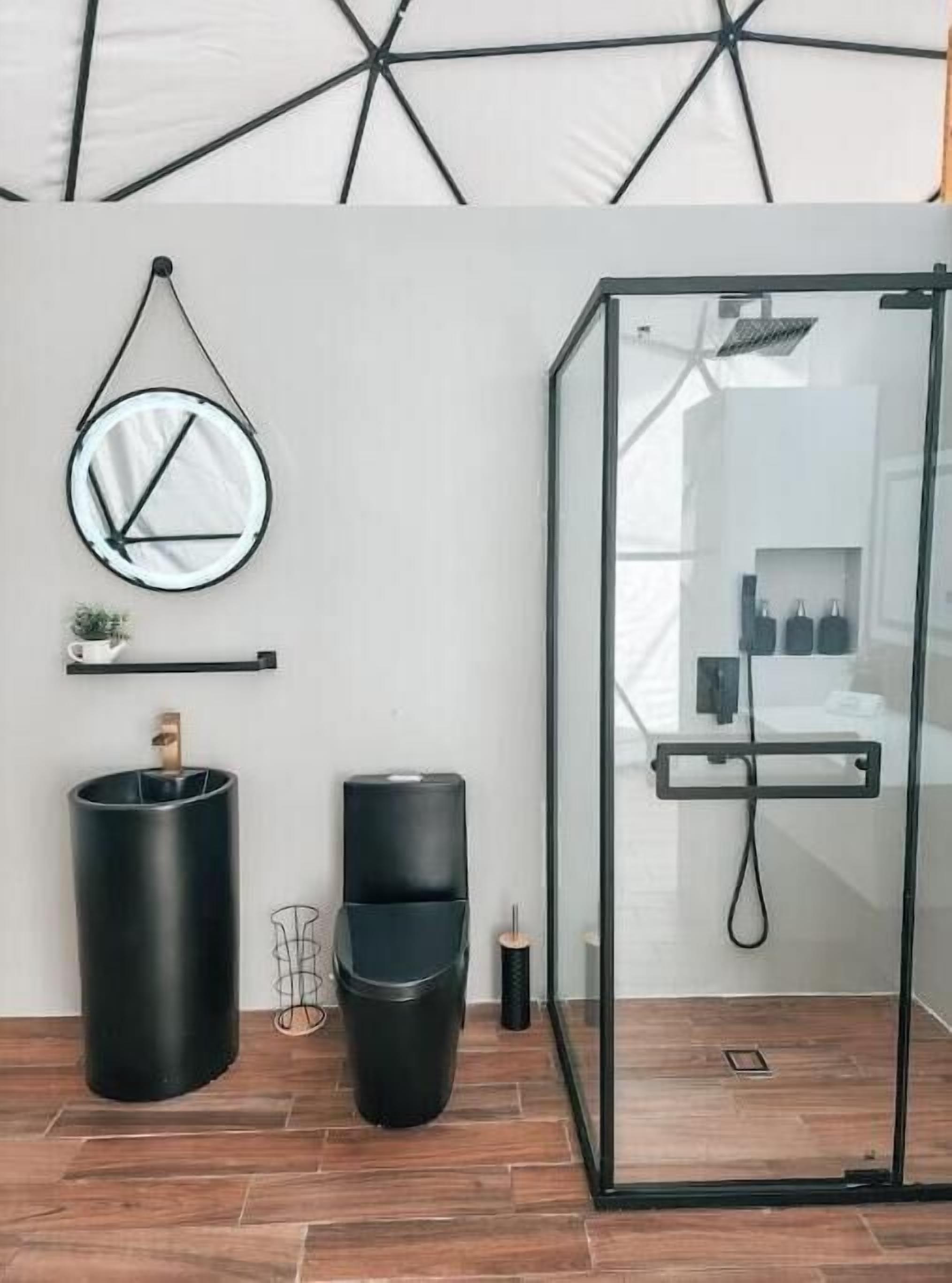 Un baño moderno con cabina de ducha de vidrio, un espejo redondo y dos recipientes cilíndricos negros en el suelo.