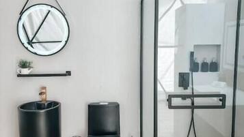 Un baño moderno con cabina de ducha de vidrio, un espejo redondo y dos recipientes cilíndricos negros en el suelo.