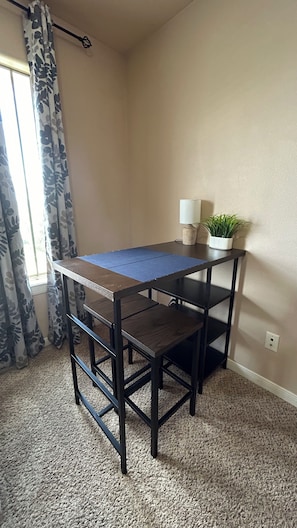 A corner room with a dark wooden table, a blue mat, a white lamp, and a potted plant.