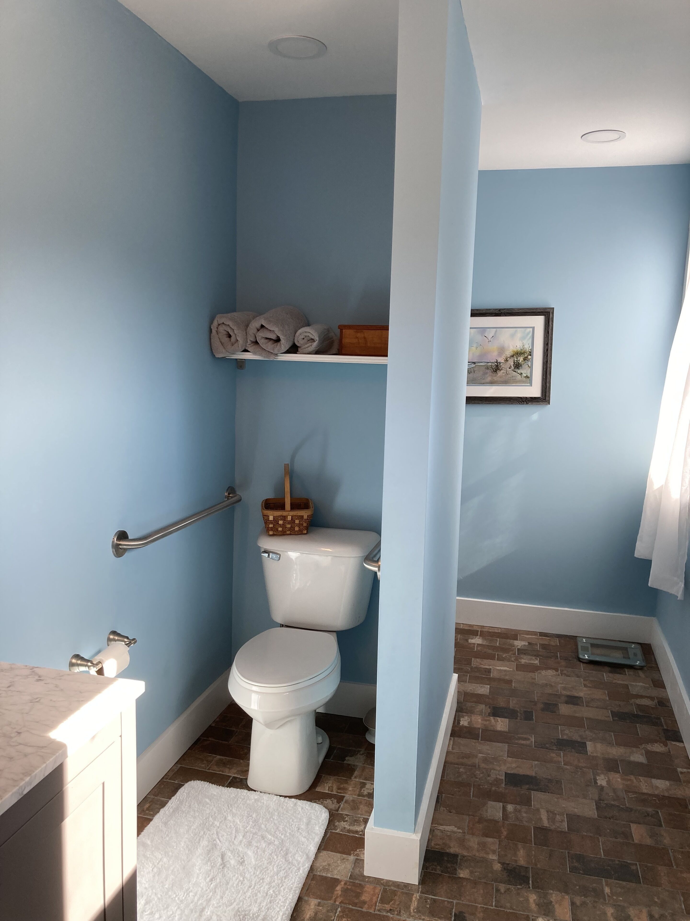 A bathroom with a toilet, a handrail, a shelf with towels, a picture on the wall, and a tiled floor.
