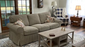A living room with a beige sectional sofa, a wooden coffee table, a patterned rug, a clock on the wall, and a view through a glass door. - Rustic and private farmhouse close to Lackawanna State Park, near Elk Mtn Ski  (Dalton)