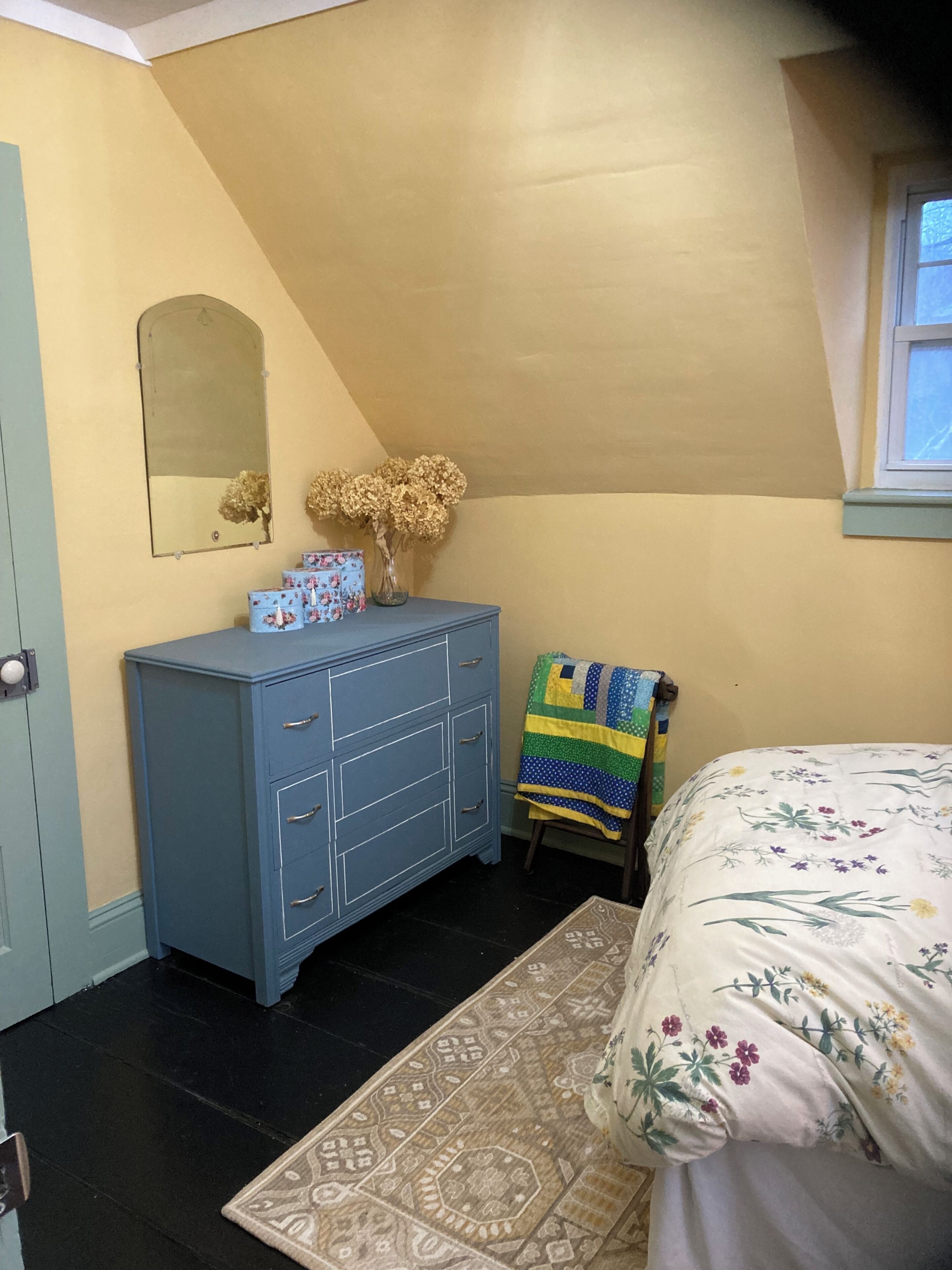 Une chambre avec une commode bleue, un miroir, un vase contenant des fleurs séchées, une couverture matelassée et un tapis à motifs.