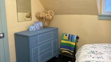 Une chambre avec une commode bleue, un miroir, un vase contenant des fleurs séchées, une couverture matelassée et un tapis à motifs.