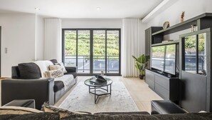 A modern living room with a sofa, coffee table, and TV. Large windows offer a view of greenery outside. - Til Home by Homie in Funchal (Funchal)