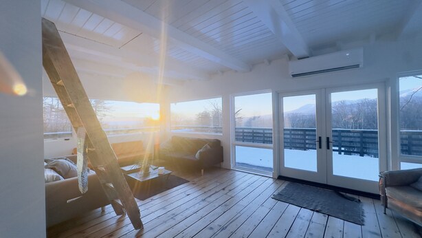 Ein sonnendurchflutetes Zimmer mit Holzboden, einer Couch, einem Sessel und Blick auf eine verschneite Landschaft durch große Glaswände.