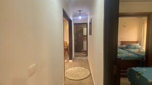 A narrow hallway with a round rug, a ceiling light fixture, and a bed with a blue bedspread in the adjacent room. - Luxurious Modern Apartment (Madinaty)