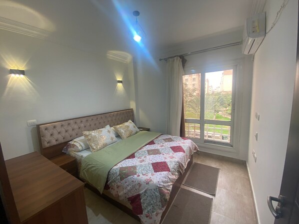 A bedroom with a bed, bedside tables, a window with a view, and a ceiling light. - Luxurious Modern Apartment (Madinaty)