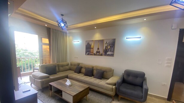 A modern living room with a sectional sofa, a coffee table, and a grey armchair. There are three wall-mounted light fixtures and a large window with curtains. - Luxurious Modern Apartment (Madinaty)